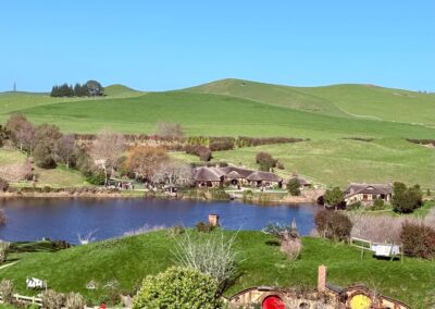 Voyage Nouvelle-Zélande en famille : Village d'hobbiton avec les trous des hobbits en premier plan, l'étang puis l'auberge. Matamata, région de Waikato.