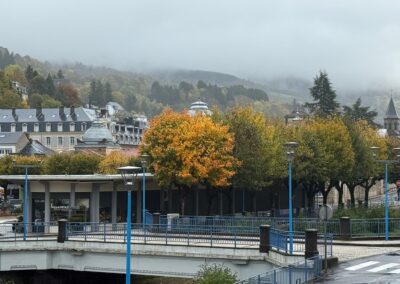 Voyage sur mesure en Auvergne : Pont aux garde-corps bleus surplombant La Bourboule, avec arbres aux couleurs d'automne et collines dans la brume