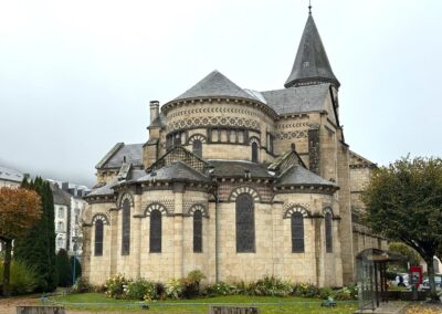 Visite hors des sentiers battus en Auvergne : Chevet roman de l’église Saint-Joseph de la Bourboule, en pierre claire avec clocher pointu, décors en mosaïque de pierre et jardin fleuri.