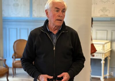 Séjour familial en Auvergne : Serge Teillot, propriétaire de Subirana s'exprime devant un groupe de visiteurs dans un salon d'époque de la Maison Rozier, tenant deux petits objets ronds dans ses mains.