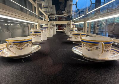 Visite hors des sentiers battus en Auvergne : Rangées de tasses, soucoupes et pots à lait en porcelaine ancienne à liseré bleu et or, exposés dans une vitrine.