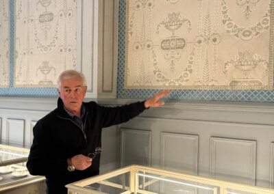 Escapade entre amis en Auvergne : Serge Teillot, propriétaire de Subirana présente un papier peint ancien à motifs néoclassiques, à côté de vitrines d'exposition.