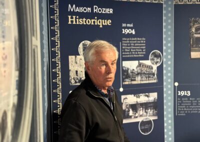 Visite hors des sentiers battus en Auvergne : Serge Teillot se tient devant un panneau bleu marine intitulé "Maison Rozier Historique", illustré de photographies d'archives datées de 1904.