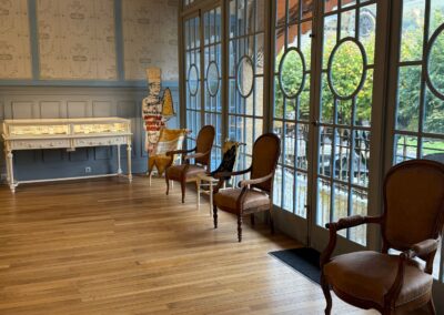 Visite hors des sentiers battus en Auvergne : Salon lumineux avec parquet en bois, fauteuils en cuir d'époque, vitrine ancienne et grandes fenêtres à petits carreaux donnant sur le parc.