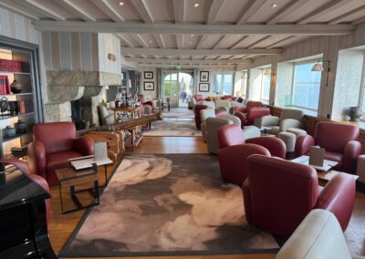 Séjour luxe familial en Bretagne : Le bar du domaine de Rochevilaine avec sa cheminée en granit, ses fauteuils en cuir rouge et ses baies vitrées ouvertes sur la mer.