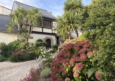 Voyage sur mesure en Bretagne : Hortensias rouges et cordylines devant une bâtisse bretonne à colombages et porte en granit au domaine de Rochevilaine.